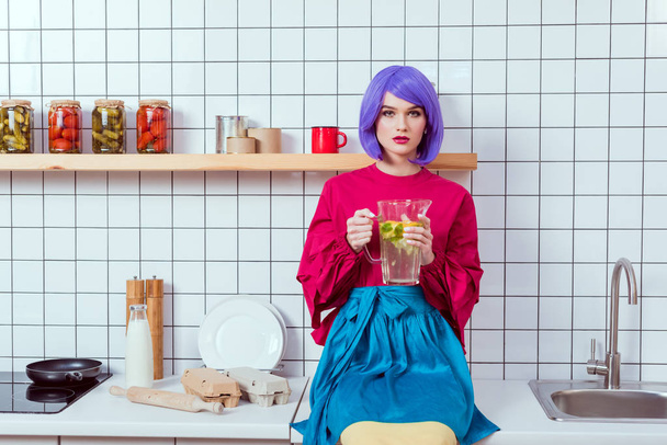 housewife with purple hair and colorful clothes sitting on kitchen counter and holding glass jar of lemonade - Foto, afbeelding