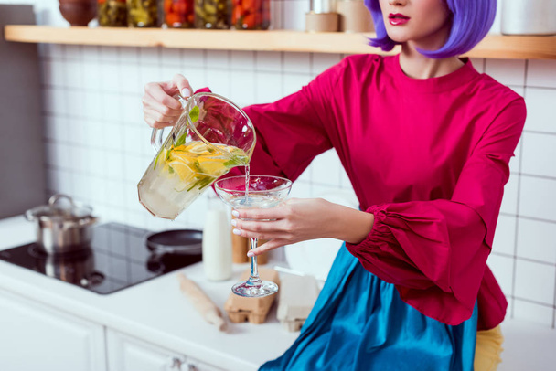 oříznutý pohled hospodyně s fialovými vlasy a barevné oblečení sedí na kuchyňské lince a nalévání limonády ve skle - Fotografie, Obrázek