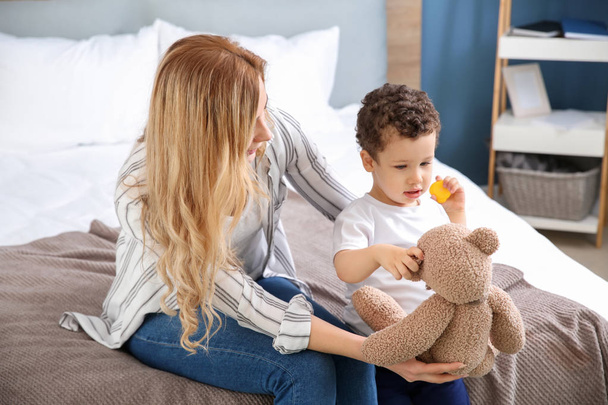 Jonge moeder met schattige kleine zoon en teddy beer zittend op bed thuis - Foto, afbeelding