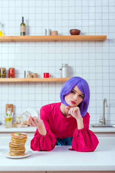 housewife with purple hair and colorful clothes pouring syrup on pancakes while looking at camera in kitchen - Photo, Image