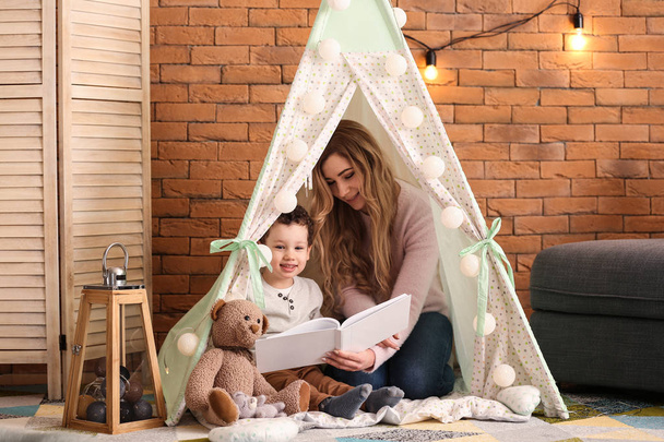 Νεαρή μητέρα με χαριτωμένο μικρό βιβλίο ανάγνωση γιο σε μια καλύβα στο σπίτι - Φωτογραφία, εικόνα
