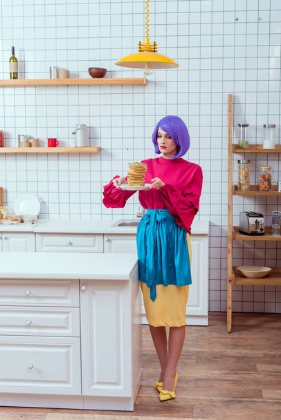 beautiful housewife in colorful clothes with purple hair holding plate with pancakes in kitchen - Foto, afbeelding