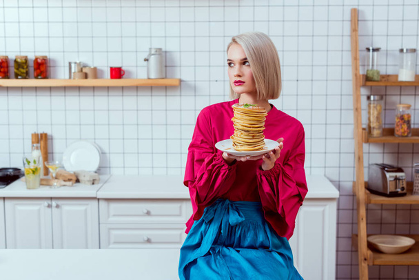 fashionable woman in colorful clothes sitting and holding plate of pancakes in kitchen - Φωτογραφία, εικόνα