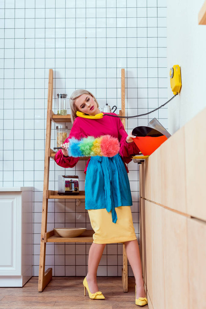 schöne Hausfrau in bunten Kleidern hält Staubbürste und spricht am Retro-Telefon in der Küche - Foto, Bild