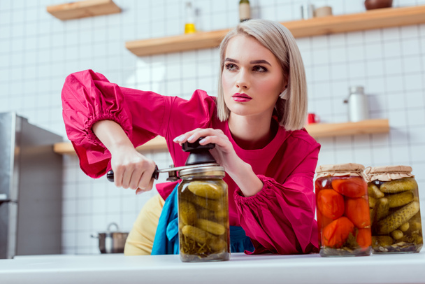 selective focus of beautiful fashionable housewife sealing jar of pickled cucumbers in kitchen  - Photo, Image