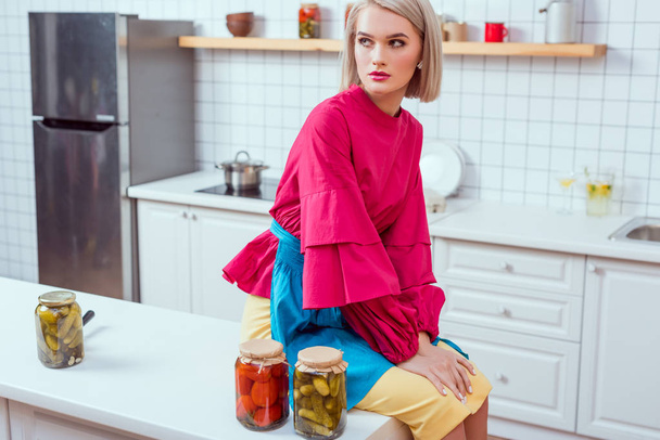 schöne modische Hausfrau sitzt auf der Küchentheke mit Gläschen mit eingelegtem Gemüse in der Küche  - Foto, Bild