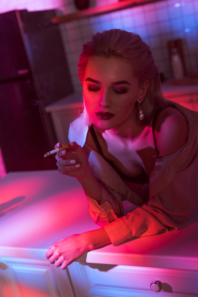 selective focus of beautiful elegant woman leaning over kitchen counter and smoking cigarette in neon pink light - Foto, Bild