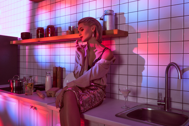 enfoque selectivo de la hermosa mujer de moda sentado en el mostrador de la cocina y fumar en luz de neón
 - Foto, Imagen