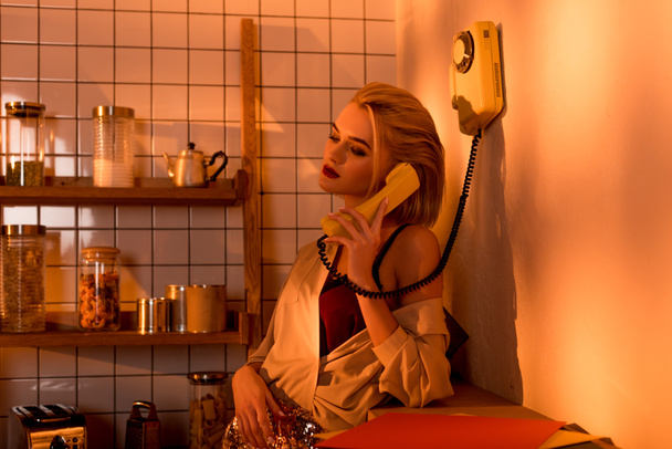 beautiful elegant young woman talking on retro telephone in kitchen with orange light - Photo, Image