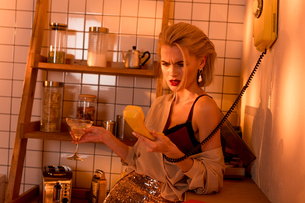 mujer frustrada sosteniendo teléfono retro y copa de cóctel en la cocina con luz naranja
 - Foto, Imagen