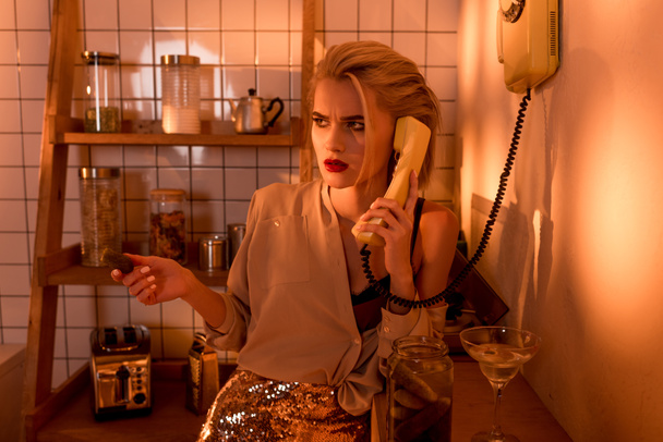 beautiful confused woman talking on retro telephone in kitchen with orange light - Photo, Image