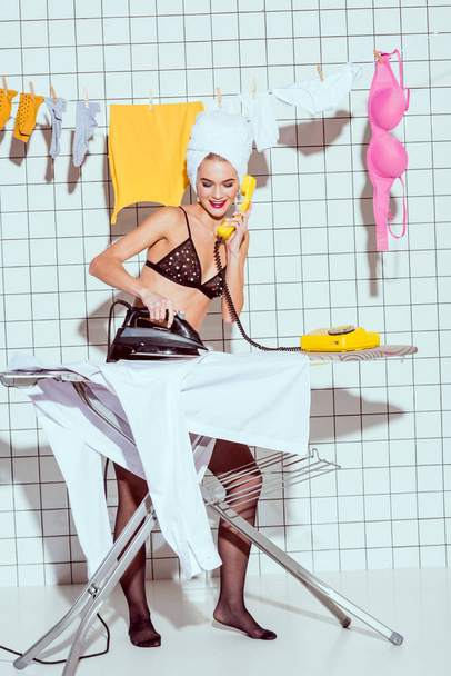 beautiful smiling housewife with towel on head ironing clothes and talking on retro telephone in bathroom - Foto, Imagem