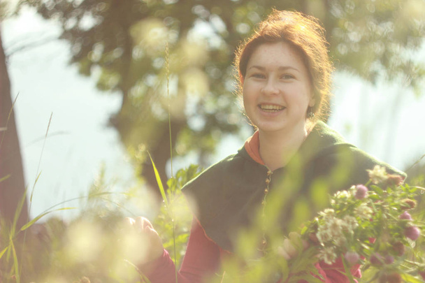 Eine fröhliche junge kaukasische weiße Frau mit langen roten Haaren lächelt und lacht mit einem Blumenstrauß in der Hand auf einem Feld voller Blumen und Sonnenlicht. heller Frühling - Foto, Bild
