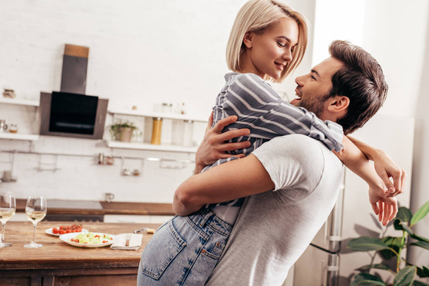 handsome boyfriend and attractive girlfriend hugging and smiling in kitchen - Foto, Imagem