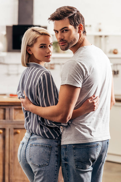 selective focus of handsome boyfriend and attractive girlfriend hugging and standing in kitchen while looking at camera - Φωτογραφία, εικόνα