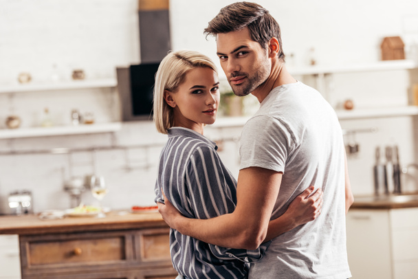 selective focus of handsome boyfriend and attractive girlfriend hugging and standing in kitchen while looking at camera - Foto, imagen