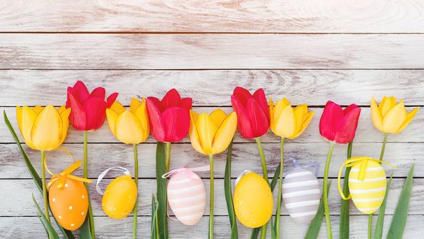 Easter eggs and tulips. Happy Easter card with copy space. Colorful easter eggs among fresh spring tulips. Decorated eggs on rustic wooden background. Easter holidays. Red and yellow spring tulips - Photo, Image