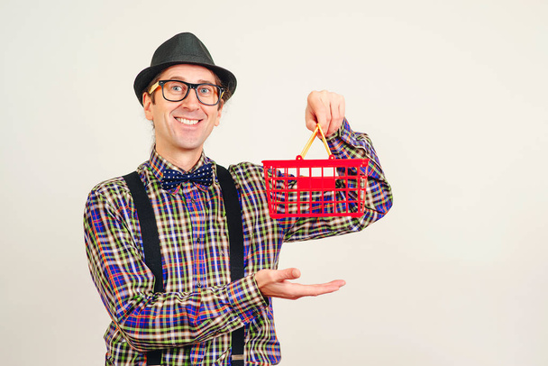 Hombre divertido en sombrero y camisa a cuadros sosteniendo cesta de supermercado rojo. Hombre de compras, aislado en blanco. Copiar espacio. Ventas estacionales o festivas. Compras en línea. Nerd en gafas con cesta de la compra
 - Foto, imagen