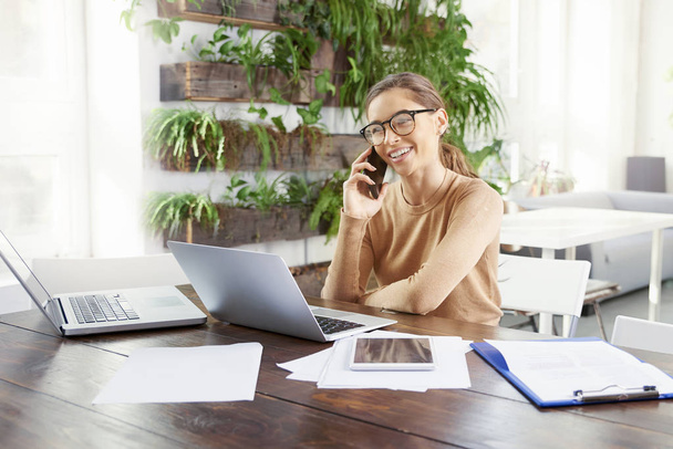 Retrato de bela jovem empresária sentada atrás de seu laptop na mesa de escritório e conversando com alguém no telefone celular enquanto trabalhava no escritório
.  - Foto, Imagem