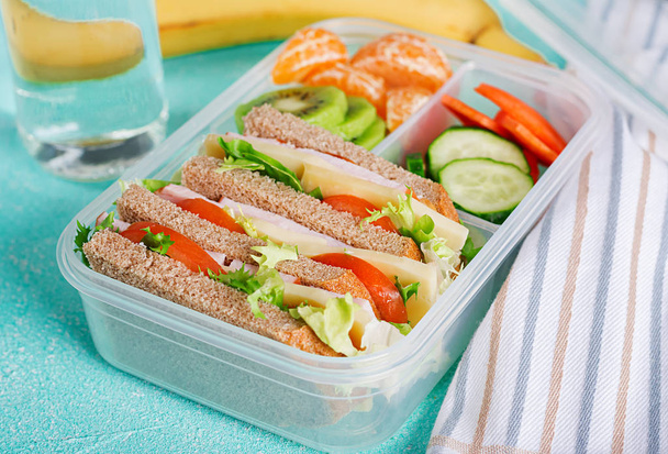 Iskolai ebéd doboz szendvicsekkel, zöldségekkel, vízzel és gyümölccsel az asztalnál. Az egészséges étkezési szokások fogalma. - Fotó, kép