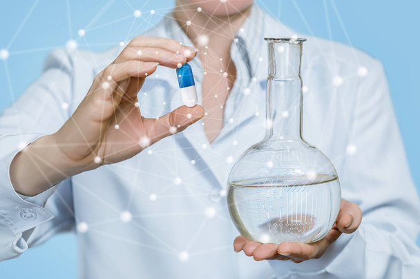 A laboratorian assistant is holding a transparent flask with substance and a medical pill in her hands at light blue background. The concept of innovative science and technology in modern medicine. - Photo, Image