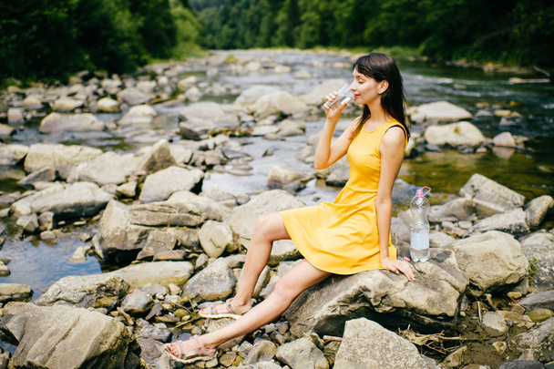 young woman drinking water at river bank with stones on shore - Foto, afbeelding