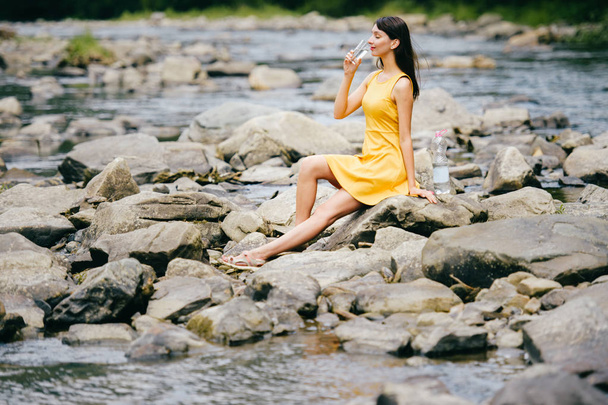 young woman drinking water at river bank with stones on shore - Foto, afbeelding