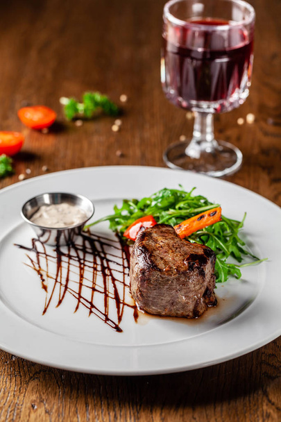 Cuisine géorgienne. Steak de bœuf juteux, steak de veau sur une assiette blanche avec roquette rôtie, légumes grillés et sauce. Sur la table se trouve un verre de vin de grenade rouge. Servant des plats au restaurant
 - Photo, image