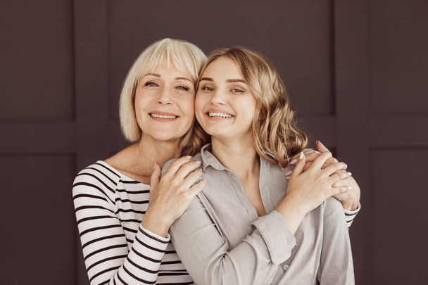 Senior woman and young daughter hugging against brown wall - Photo, Image