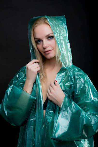 Portrait of a young blond woman in a green raincoat looking at the camera, a hood is put on her head. Shooting in a photo studio on a black background - Фото, изображение