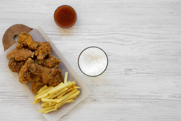 Fast food: fried chicken legs, spicy wings, French fries and chicken tenders with sour-sweet sauce and cold beer over white wooden background, overhead view. Flat lay, overhead, from above. Copy space. - Photo, Image