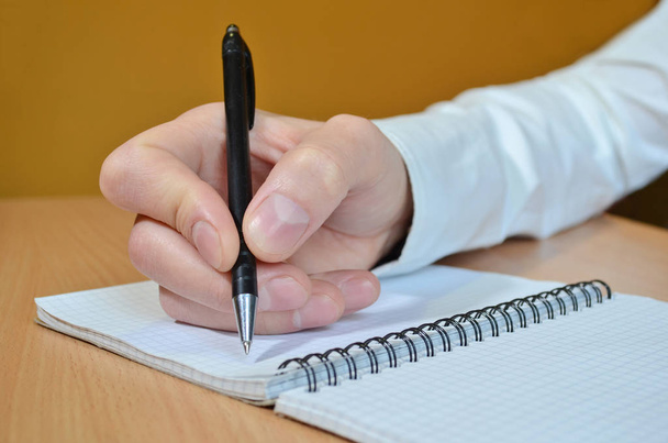 Мужская рука в белой рубашке пишет текст черной ручкой в блокноте со спиралью на деревянном столе
 - Фото, изображение