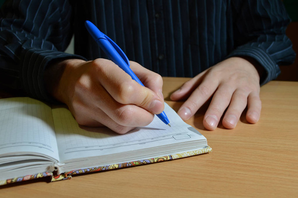 Мужская рука в синей рубашке пишет текст черной ручкой в блокноте на деревянном столе, другой рукой
 - Фото, изображение