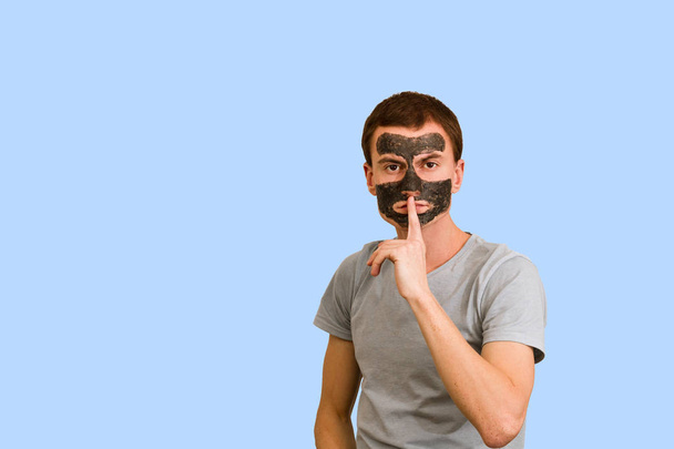 Guy in a black cosmetic mask on a blue background. Facial skincare. Concept of facial problems - 写真・画像