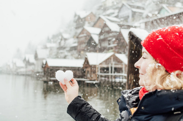Young blonde smiling woman in a red hat holding snow ball in a shape of a heart in front of the old village of Hallstatt, Austria. Blurry background, shallow depth of field. - Photo, Image