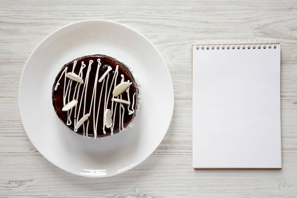Homemade chocolate cherry cake and blank notepad on white wooden background, view from above. Flat lay, top view. Space for text. - Foto, imagen