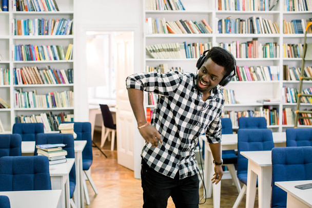 A handsome young African American man in casual clothes in a library while listening to music with headphones and dancing to the music. Concept: educational, portrait, library, dancer and music - Valokuva, kuva