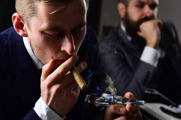 He has megabucks. Businessmen have money to burn. Man light up cigar from money banknote. Rich man smoking during business meeting. Business partners writing financial report. Waste of money - Foto, Bild