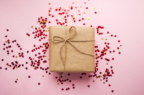 Gift box wrapped in brown colored craft paper and tied with rope on pink background with heart shape red confetti. - Photo, Image
