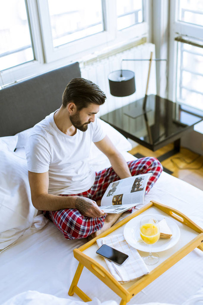 Νέοι γενειοφόρος άνδρας κατοχής του προγεύματος στο κρεβάτι και την ανάγνωση περιοδικών στο δωμάτιο - Φωτογραφία, εικόνα