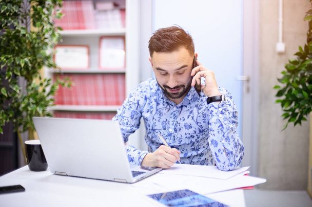 Бизнесмен работает над ноутбуком и делает телефонный звонок в своем офисе. Человек, работающий в офисе
 - Фото, изображение
