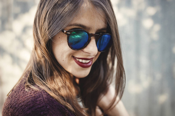 Élégante fille hipster souriant dans la rue ensoleillée sur fond de mur en bois. Boho fille en tenue cool et lunettes de soleil posant dans la lumière du soleil et l'ombre. Espace pour le texte. Vacances d'été et voyages
 - Photo, image