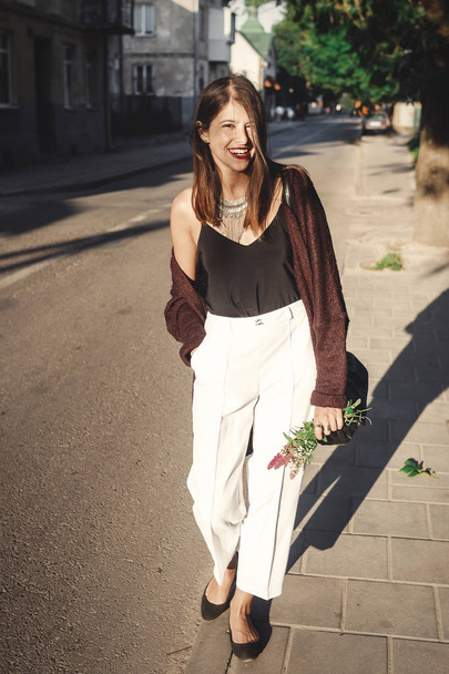 Stylish bohemian girl with modern jewelry walking in sunny street. Happy hipster carefree girl in boho look with flowers relaxing in evening summer city. Space for text. Summer vacation - Foto, Imagem