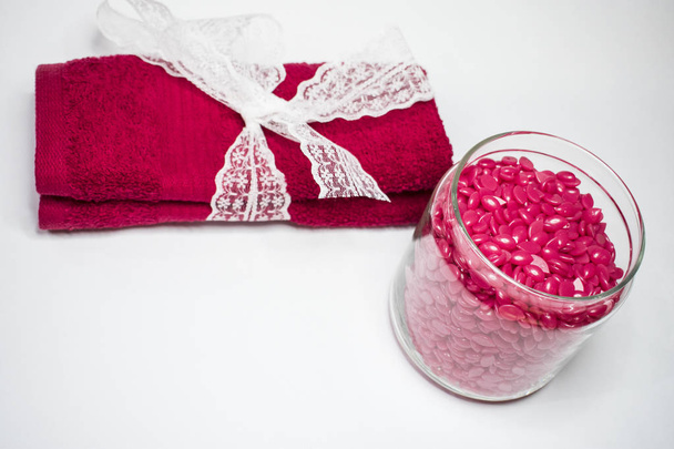 Isolado em um fundo branco é um frasco de vidro com cera granulada vermelha para depilação e uma toalha vermelha com um arco branco
. - Foto, Imagem