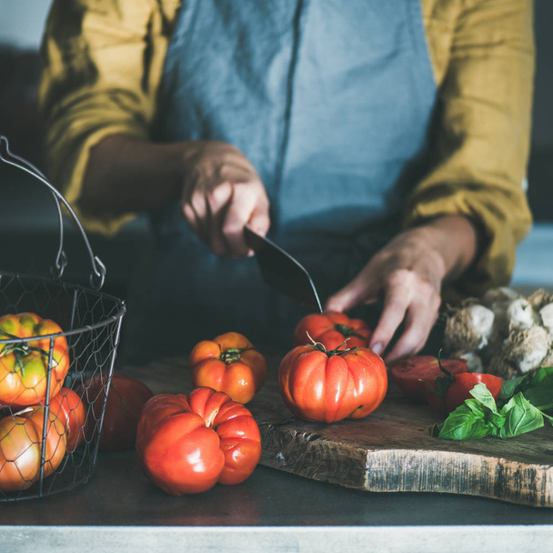 Woman in grey linen apron cooking tomato sauce, canned tomatoes or pasta with heirloom tomatoes, basil and garlic at kitchen counter, square crop. Healthy cooking, slow food or comfort food - 写真・画像