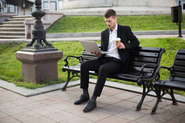 Young handsome businessman in classic black suit and white shirt with wireless earphones and cup of coffee to go in hand thoughtfully working on laptop sitting on bench outdoor - Photo, Image