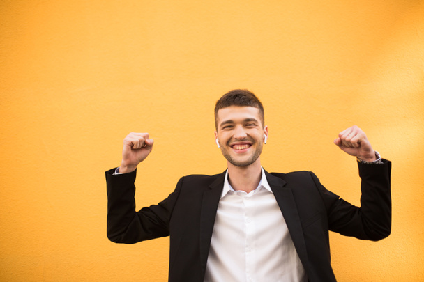 Young joyful man in classic black jacket and white shirt with wireless earphones happily showing yes gesture looking in camera over orange background - 写真・画像