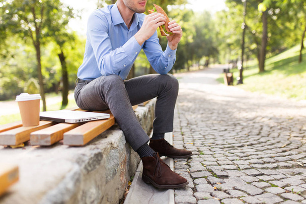 Cerca de hombre sentado en el banco sosteniendo sándwich en las manos con una taza de café para ir y portátil cerca de pasar tiempo en el acogedor parque
 - Foto, imagen