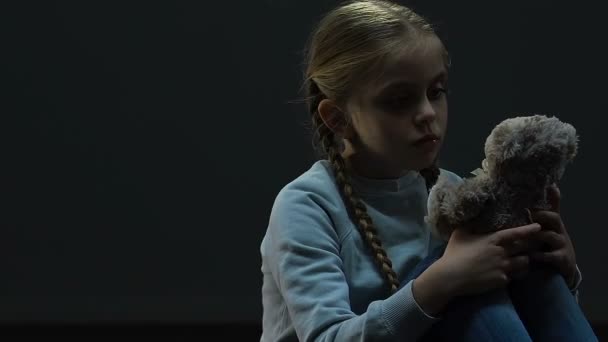 Kleine meisje knuffelen toy bear zitten in donkere lege ruimte, gebrek aan ouderlijke zorg - Video