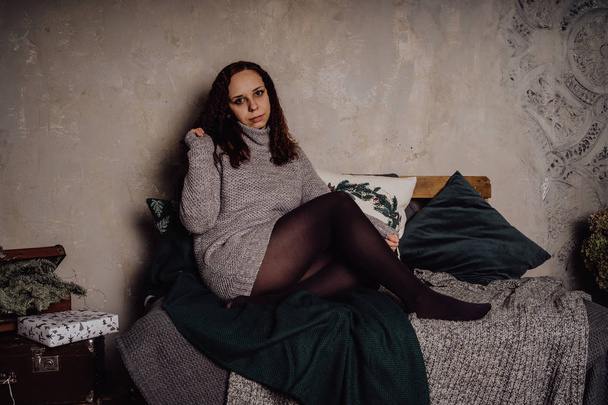 A young girl in a sweater sitting on the bed huddled in a blanket. Woman dressed in warm sweater, street style clothing. Clean skin, long hair - Valokuva, kuva
